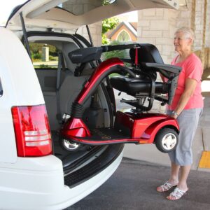 A woman stands by a white vehicle as a red mobility scooter is lifted into the cargo area using a Bruno VSL-6000 Curb-Sider® Hoist Style Interior Lift.