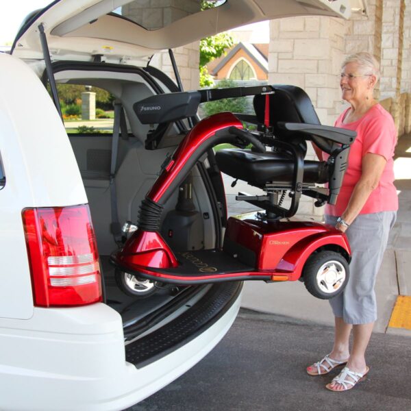 A woman stands by a white vehicle as a red mobility scooter is lifted into the cargo area using a Bruno VSL-6000 Curb-Sider® Hoist Style Interior Lift.
