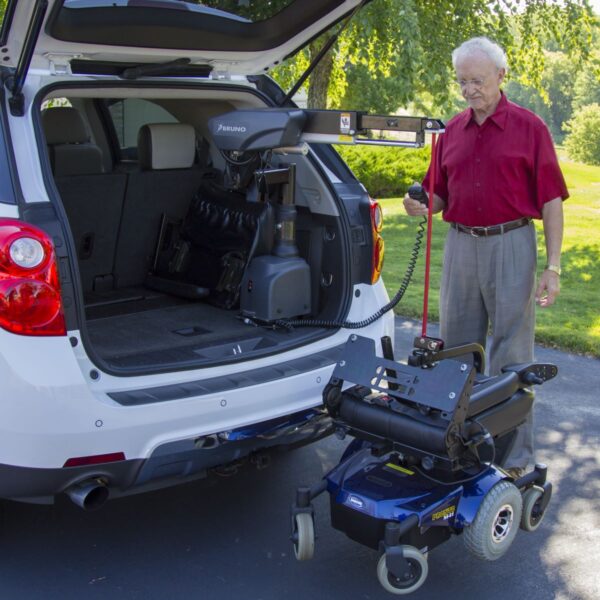 An older man uses a Bruno VSL-6000 Curb-Sider® Hoist Style Interior Lift in the back of an SUV to load an electric wheelchair on a sunny day outdoors.