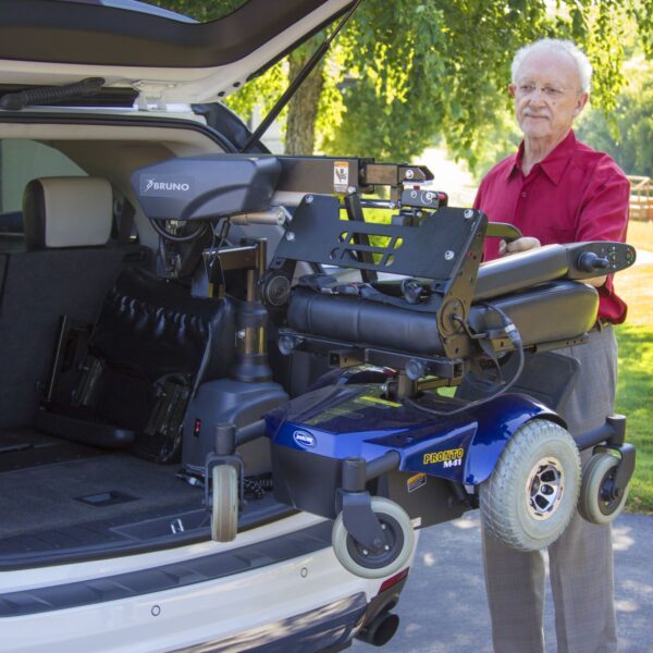 An older man stands by a vehicle with its trunk open, demonstrating the Bruno VSL-6000 Curb-Sider® Hoist Style Interior Lift as it lifts a mobility scooter.