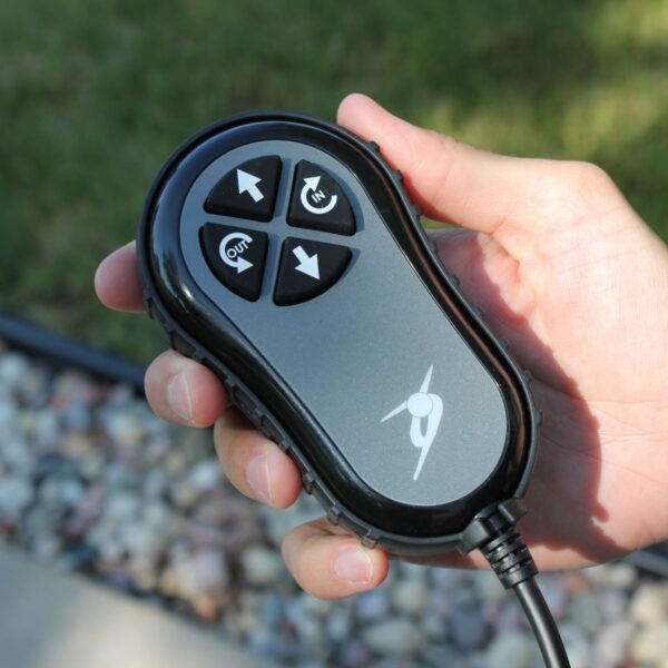 A hand holds the wired remote control of the Bruno VSL-6000 Curb-Sider® Hoist Style Interior Lift near rocks and grass, using its directional and in/out buttons to operate the device.