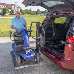 An older man stands beside a blue mobility scooter as a Bruno Joey Platform Style Interior Lift (VSL-4400) lifts it on a platform into the back of a red vehicle.