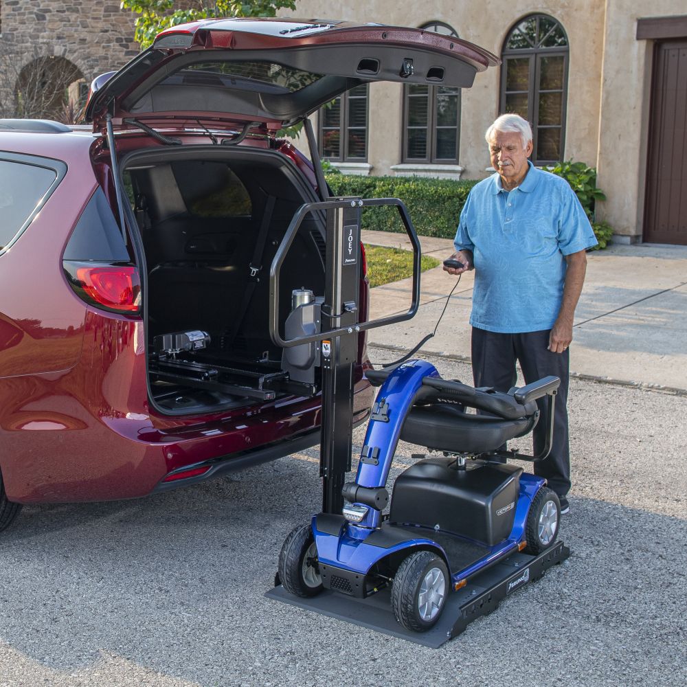 An older man uses a remote to operate a vehicle lift, loading a blue mobility scooter into the back of a red minivan parked in a driveway.
