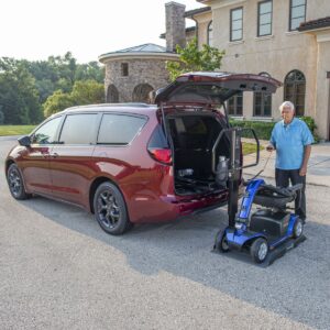 An older man stands beside a red minivan with its rear hatch open, using a Bruno Joey Platform Style Interior Lift (VSL-4400) to load a blue mobility scooter into the vehicle.