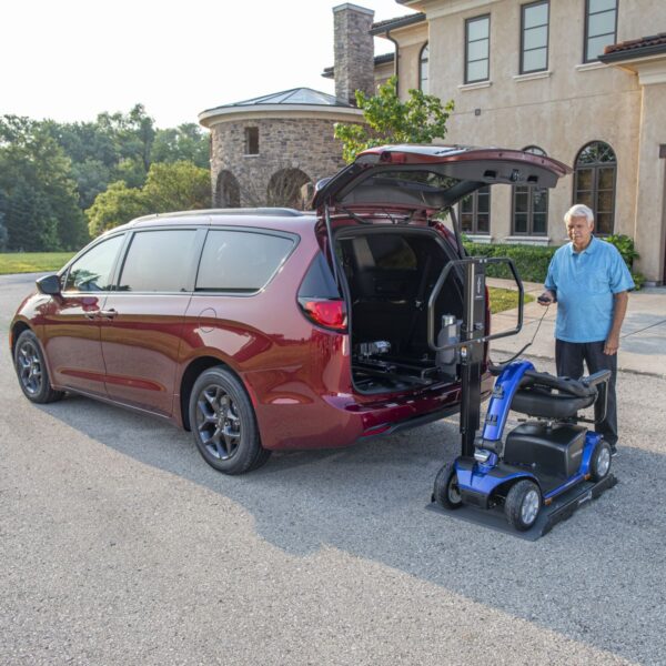 An older man stands beside a red minivan with its rear hatch open, using a Bruno Joey Platform Style Interior Lift (VSL-4400) to load a blue mobility scooter into the vehicle.