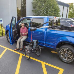 A man sits in the driver’s seat of a blue pickup parked in an accessible spot, next to a Bruno PUL-1100HD Out-Rider® wheelchair lift and an unfolded wheelchair.