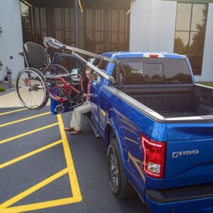 A man uses a Bruno PUL-1100HD Out-Rider®, mounted on a blue Ford F-150, to lift a wheelchair into the truck bed near a building entrance.