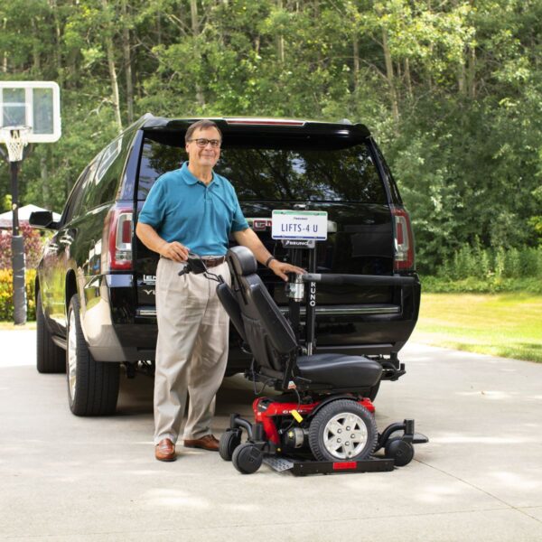 A man stands beside a black SUV with a Bruno Out-Sider® (ASL-275) scooter lift, holding his wheelchair as the lift extends from the rear. A basketball hoop and trees appear in the background.