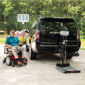 A man in a motorized wheelchair is outside next to a black SUV equipped with the Bruno Out-Sider® (ASL-275) scooter lift, featuring a rear platform. A basketball hoop appears in the background.
