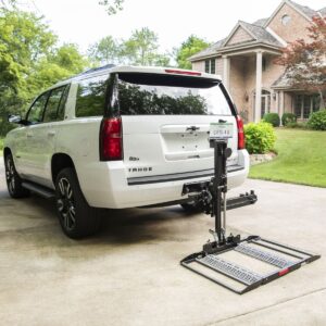 A white SUV with a Bruno Out-Sider® (ASL-275) scooter lift is parked in a residential driveway in front of a brick house.