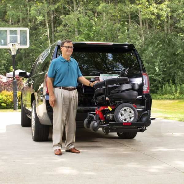 A man stands beside an SUV fitted with a Bruno Out-Sider® (ASL-275) lift carrying a mobility scooter on the back, in a tree-lined driveway with a basketball hoop visible in the background.