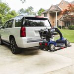 A Bruno Out-Sider® (ASL-275) scooter lift is mounted to the rear of a white Chevrolet Tahoe, carrying a mobility scooter on a platform in a residential driveway.