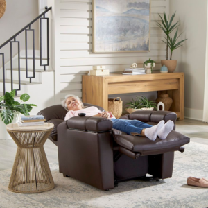 An older woman relaxes with her feet elevated in a Cloud+ Lift Chair, surrounded by plants, a wooden console table, and wall art in the living room.
