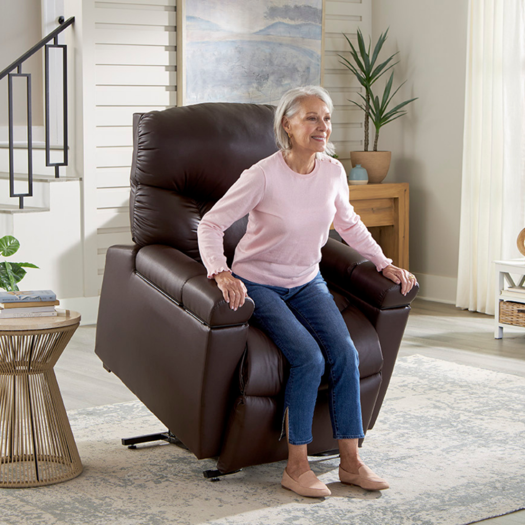 An older woman in a pink sweater stands up from the brown Cloud+ Lift Chair in a bright, modern living room.