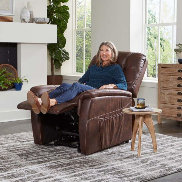 A woman smiles and reclines in the Dione Lift Chair, a brown leather armchair, in a bright living room with her feet up and a drink on a small wooden table nearby.