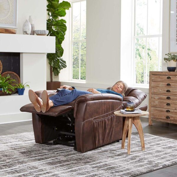 A person reclines with eyes closed on the Dione Lift Chair—a brown leather chair in a bright living room, with a drink on a small table nearby.