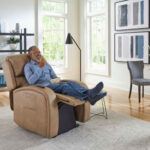 A man sits in a tan EZ Sleeper Slim with Twilight Lift Chair with his feet up, eating popcorn and watching TV in a bright, modern living room.