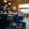 A man in a Jazzy® Ultra Light Travel Power Wheelchair receives a drink from a barista at a café counter, with the menu visible in the background.