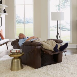 An older man relaxes in the Rhea Lift Chair, his eyes closed and hands on his chest, in a bright living room featuring a side table, lamp, and large windows.
