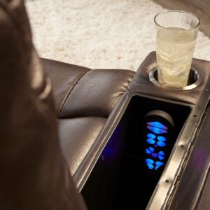 A glass with a light beverage rests in the cup holder of the Rhea Lift Chairs brown leather armrest, beside a compartment holding a remote control with illuminated buttons.