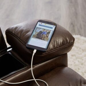 A smartphone charges on the armrest of the Rhea Lift Chair, a brown leather recliner; a website is displayed on the screen and part of a carpet can be seen in the background.