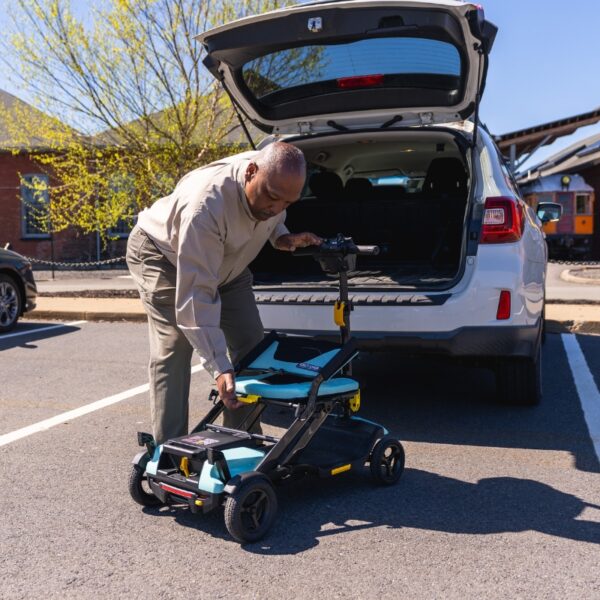 A man folds a Go Go® Super Portable 4-Wheel Scooter beside the open trunk of a parked SUV in a sunny parking lot.