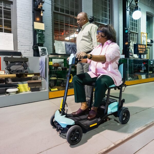 Two people are observing a display; one is sitting on a Go Go® Super Portable 4-Wheel Scooter while the other stands beside them. They are indoors near a glass case showcasing various objects.